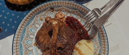 Ein Tisch mit einem Teller Lamm, Kartoffelpüree, Sauce und einer kleinen Kürbis. Ein Weinglas, eine Tasse und eine Kerze stehen ebenfalls auf dem Tisch.