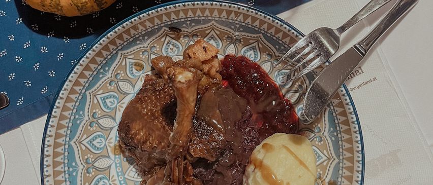 Ein Tisch mit einem Teller Lamm, Kartoffelpüree, Sauce und einer kleinen Kürbis. Ein Weinglas, eine Tasse und eine Kerze stehen ebenfalls auf dem Tisch.