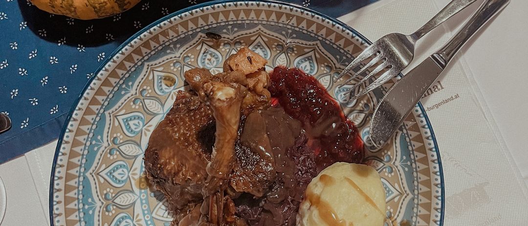 Ein Tisch mit einem Teller Lamm, Kartoffelpüree, Sauce und einer kleinen Kürbis. Ein Weinglas, eine Tasse und eine Kerze stehen ebenfalls auf dem Tisch.