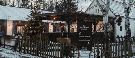 Ein Museum mit einem Schild, das 'dazumal' besagt, ist schneebedeckt. Vor dem Gebäude steht ein Weihnachtsbaum, und ein Zaun umgibt den Vorgarten. Vor dem Gebäude stehen zwei Räder.