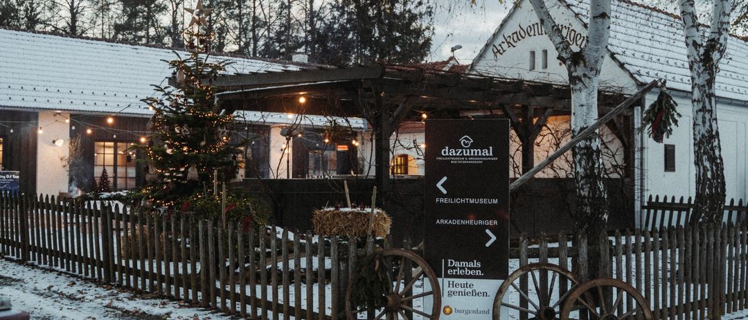 Ein Museum mit einem Schild, das 'dazumal' besagt, ist schneebedeckt. Vor dem Gebäude steht ein Weihnachtsbaum, und ein Zaun umgibt den Vorgarten. Vor dem Gebäude stehen zwei Räder.