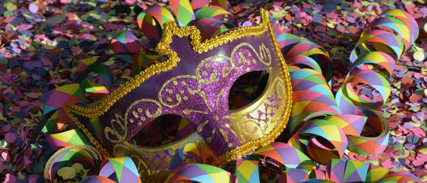 Eine lila und goldene Maske mit aufwendigen Verzierungen liegt auf einem Bett aus bunten Konfetti und Bändern. Die Maske zeichnet sich durch goldene Verzierungen und ein dekoratives Muster aus.