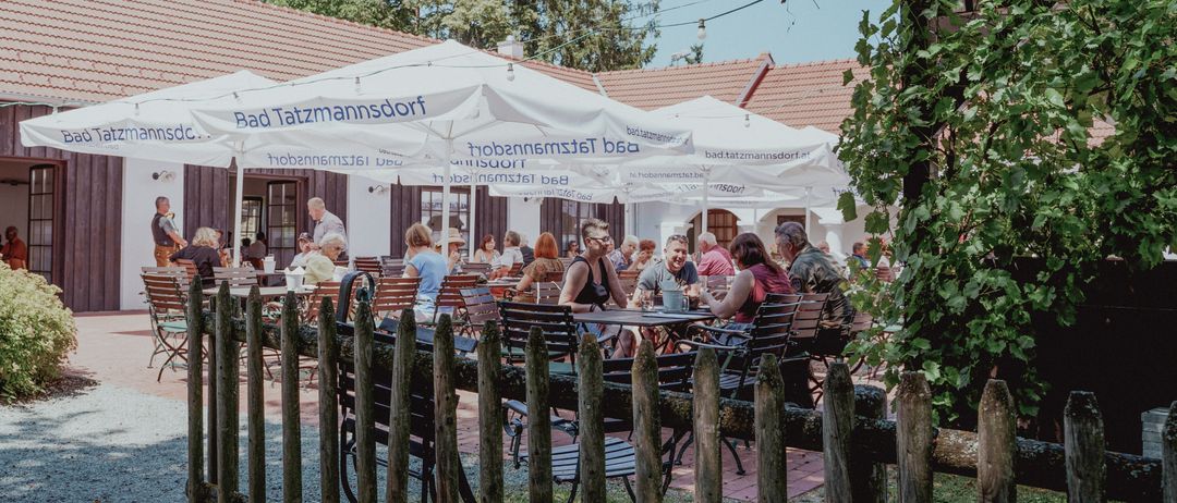 Ein Gasthof mit mehreren Gästen, die an Tischen unter Sonnenschirmen sitzen. Das Gebäude dahinter trägt ein Schild mit der Aufschrift 'Bad Tatzmannsdorf'. Bäume und Grünpflanzen sind sichtbar.
