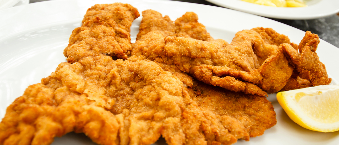 Nahaufnahme von gebratenem Schnitzel auf einem weißen Teller, goldbraun und knusprig, mit einer Zitronenscheibe in der Nähe. Im Hintergrund eine Schüssel mit cremigem Kartoffelsalat.