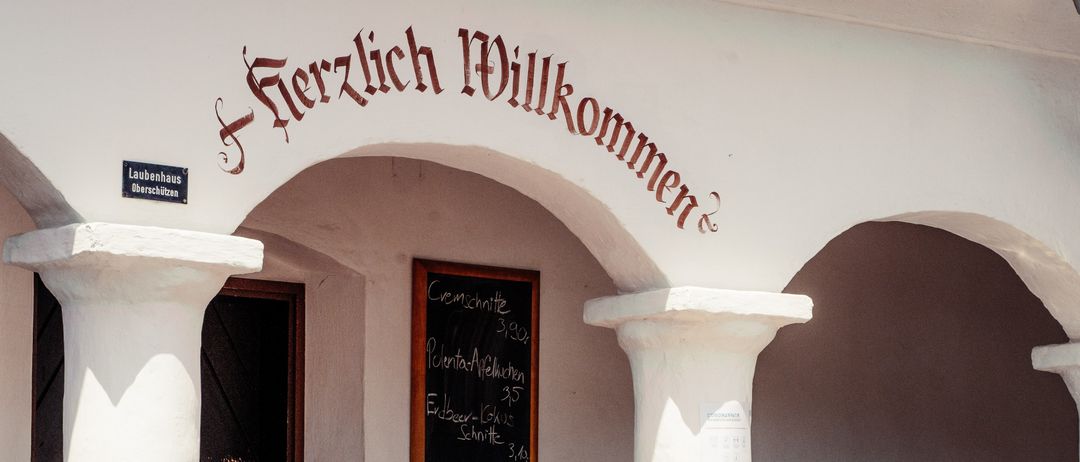 Ein Restauranteingang mit einem Schild, das 'Herzlich Willkommen' besagt. Eine schwarze Tafel mit dem Menü ist an der Wand montiert, und ein Tisch ist mit Brot, Gemüse und anderen Speisen gedeckt.