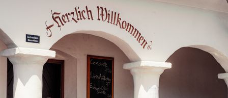 A restaurant entrance with a sign reading 'Herzlich Willkommen'. A blackboard menu is mounted on the wall, and a table is set with bread, vegetables, and other food items.