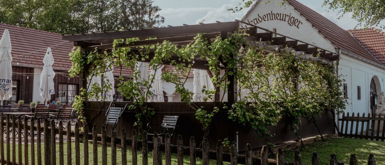 Ein rustikales Restaurant mit rotem Dach und grünen Weinranken. Ein Holzzaun umgibt den Außenbereich. Mehrere geschlossene weiße Sonnenschirme stehen neben den Tischen. Bäume und Pflanzen umgeben das Gelände.