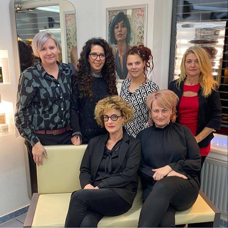 Sechs Frauen stehen in einem Raum, drei auf einer Bank und drei im Stehen. Die sitzenden Frauen tragen eine Brille und schwarze Kleidung, während die stehenden Frauen verschiedene Farben tragen. Ein Poster einer Frau mit grünen Haaren hängt an der Wand hinter ihnen.