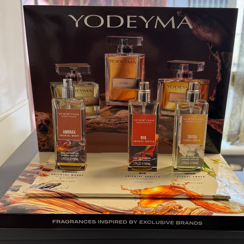 Fünf Parfümflaschen, die mit Ambrax, Nya, Tayda und zwei Yodeyma beschriftet sind, werden auf einem Tisch mit einem hölzernen Hintergrund ausgestellt. Die Yodeyma-Flaschen sind größer.