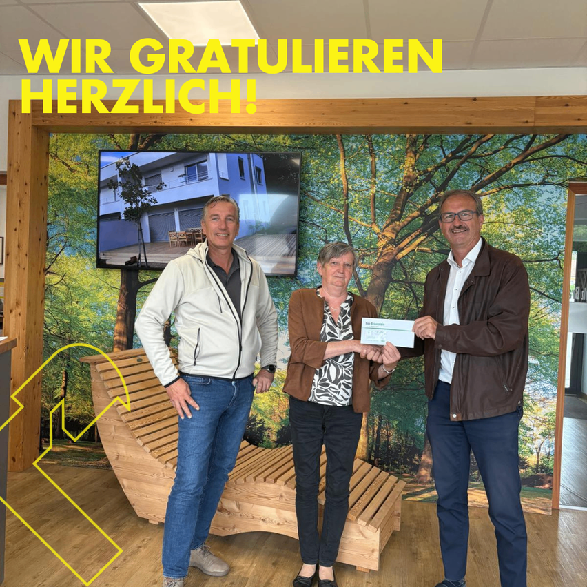 Drei Menschen stehen vor einer Wand mit einem Bild eines Hauses und lächeln. Sie halten ein Zertifikat in der Hand. Der Text darüber lautet 'Wir Gratulieren Herzlich!'. Eine Holzbank befindet sich links. Der Boden hat eine gelbe Linie.