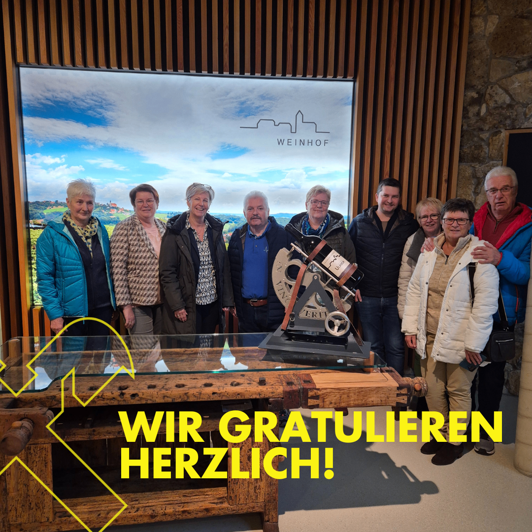 Eine Gruppe von Menschen steht vor einem Glastisch mit einem Pokal. Hinter ihnen ist eine große Leinwand mit einer Landschaftsansicht und dem Wort Weinhof. Sie alle lächeln und scheinen für ein Foto zu posieren.