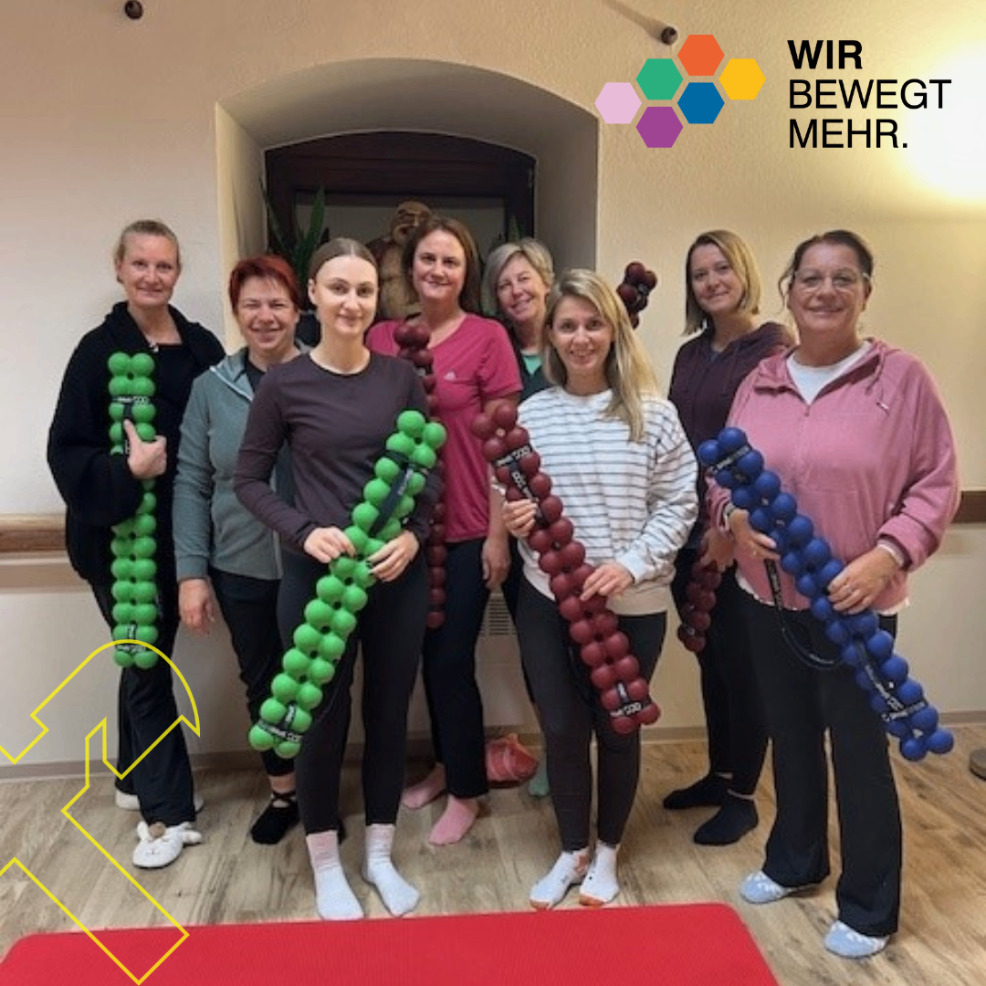 Gruppe von Frauen, die lächelnd für ein Foto posieren und farbige Ballongewichte halten, mit Textüberlagerung 'Wir bewegen uns mehr.'