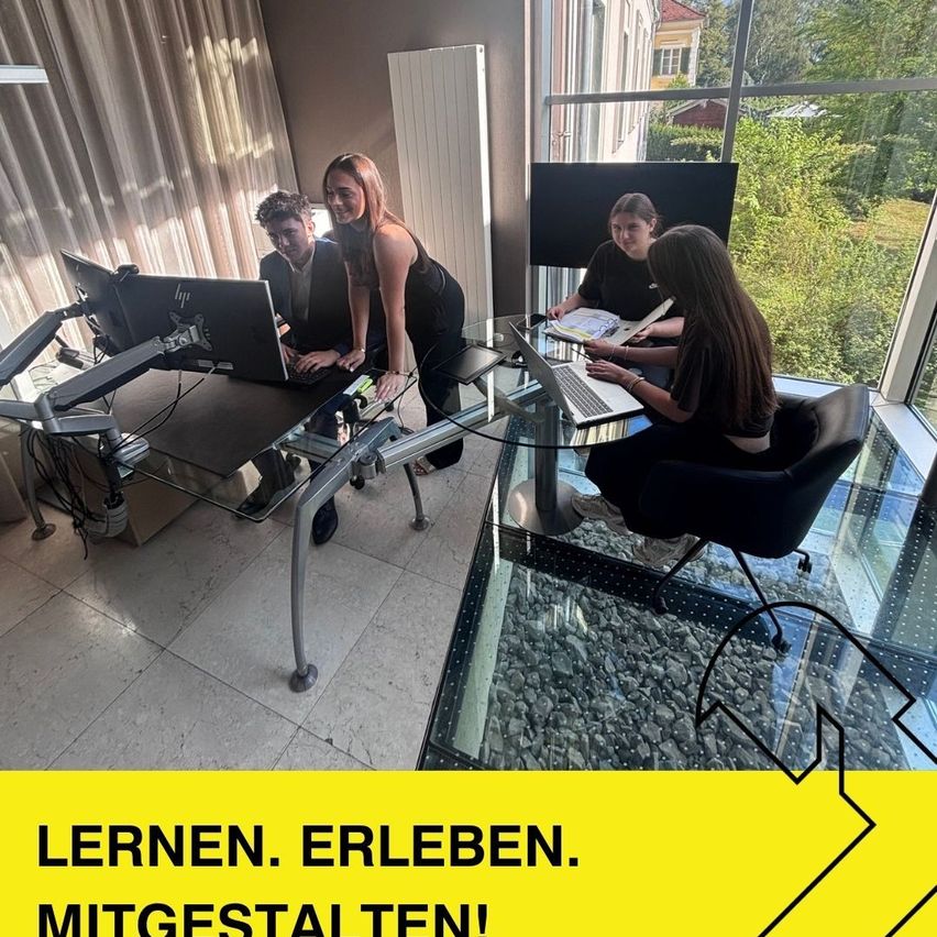 Vier Personen arbeiten in einem Büro mit Glaswänden, die einen Blick auf einen Garten ermöglichen. Zwei Frauen sitzen an einem Schreibtisch, eine tippt auf einem Laptop, während zwei Männer hinter ihnen stehen, einer lehnt sich über einen Monitor.