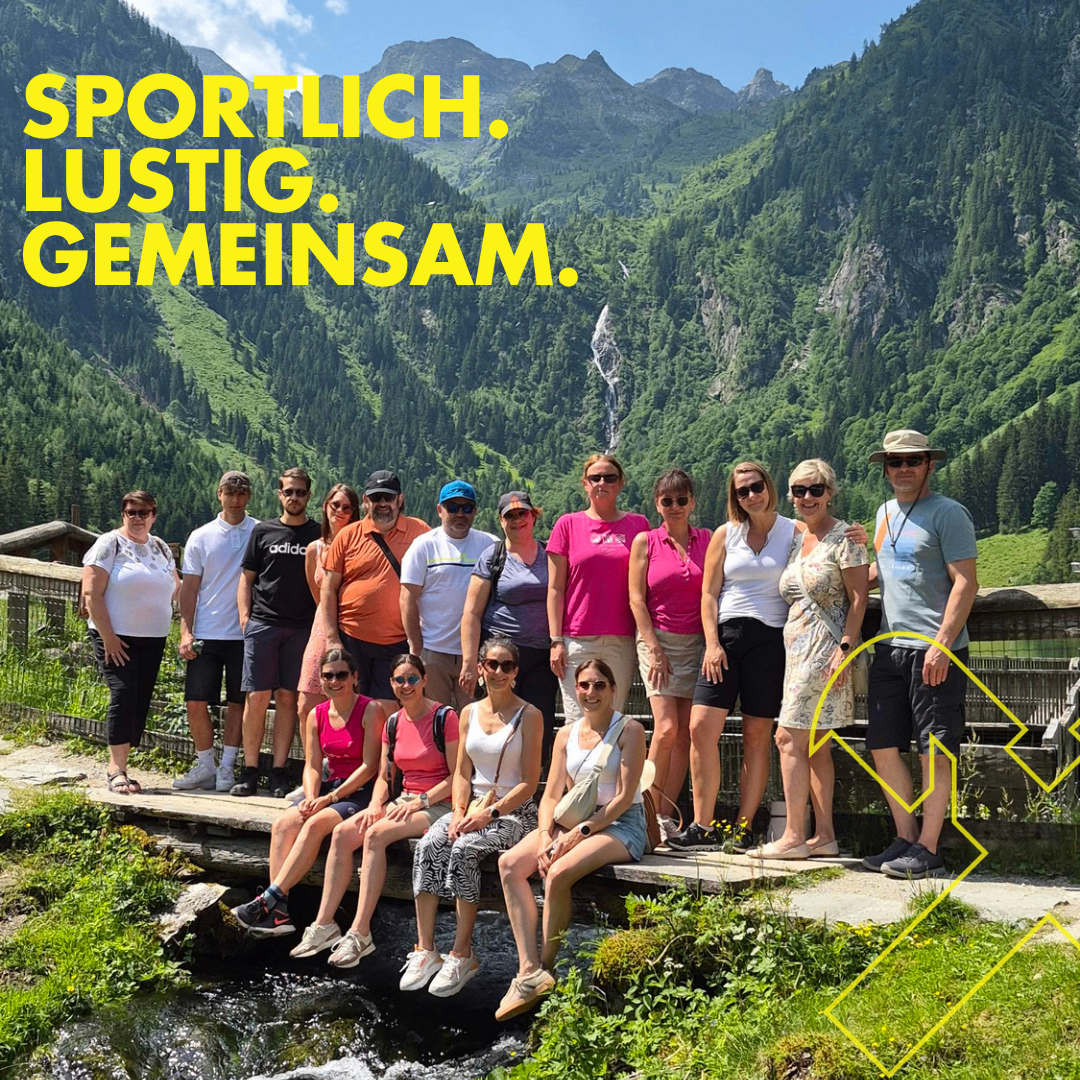 Eine Gruppe von Menschen in leuchtender Outdoor-Kleidung posiert für ein Foto an einem Bach mit Bergen im Hintergrund. Die Worte 'Sportlich, Lustig, Gemeinsam' sind in fettem gelbem Text angezeigt.