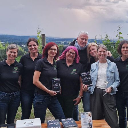 Eine Gruppe von Menschen, einige mit Büchern in der Hand, steht gemeinsam in einem Weinberg. Sie lächeln und posieren für ein Foto. Im Hintergrund ist ein bewölkter Himmel und eine entfernte Landschaft zu sehen.