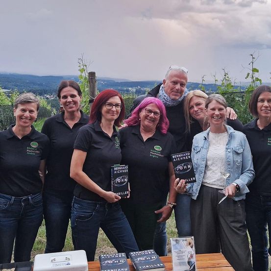 Eine Gruppe von Menschen, einige mit Büchern in der Hand, steht gemeinsam in einem Weinberg. Sie lächeln und posieren für ein Foto. Im Hintergrund ist ein bewölkter Himmel und eine entfernte Landschaft zu sehen.