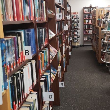 Ein Bibliotheksgang mit hölzernen Bücherregalen, die mit A bis G beschriftet sind und mit verschiedenen Büchern gefüllt sind. Der Boden ist mit Teppich ausgelegt, und im Hintergrund befinden sich zusätzliche Regale und ein Wagen.
