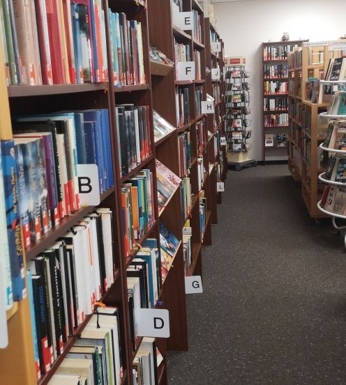Ein Bibliotheksgang mit hölzernen Bücherregalen, die mit A bis G beschriftet sind und mit verschiedenen Büchern gefüllt sind. Der Boden ist mit Teppich ausgelegt, und im Hintergrund befinden sich zusätzliche Regale und ein Wagen.