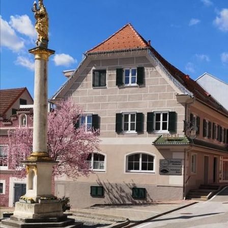Eine Statue auf einer Säule steht vor einem traditionellen Gebäude mit rotem Dach und grünen Fensterläden. Das Gebäude ist von einer Straße und rosa Bäumen umgeben.