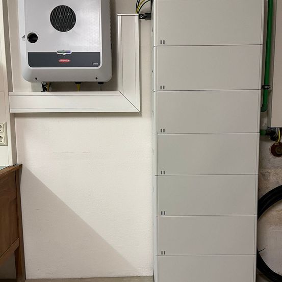 Ein weißes Wandgerät zur Stromversorgung ist an einer weißen Wand installiert, neben einem Stromzähler. Ein weißer Metallschrank steht daneben.