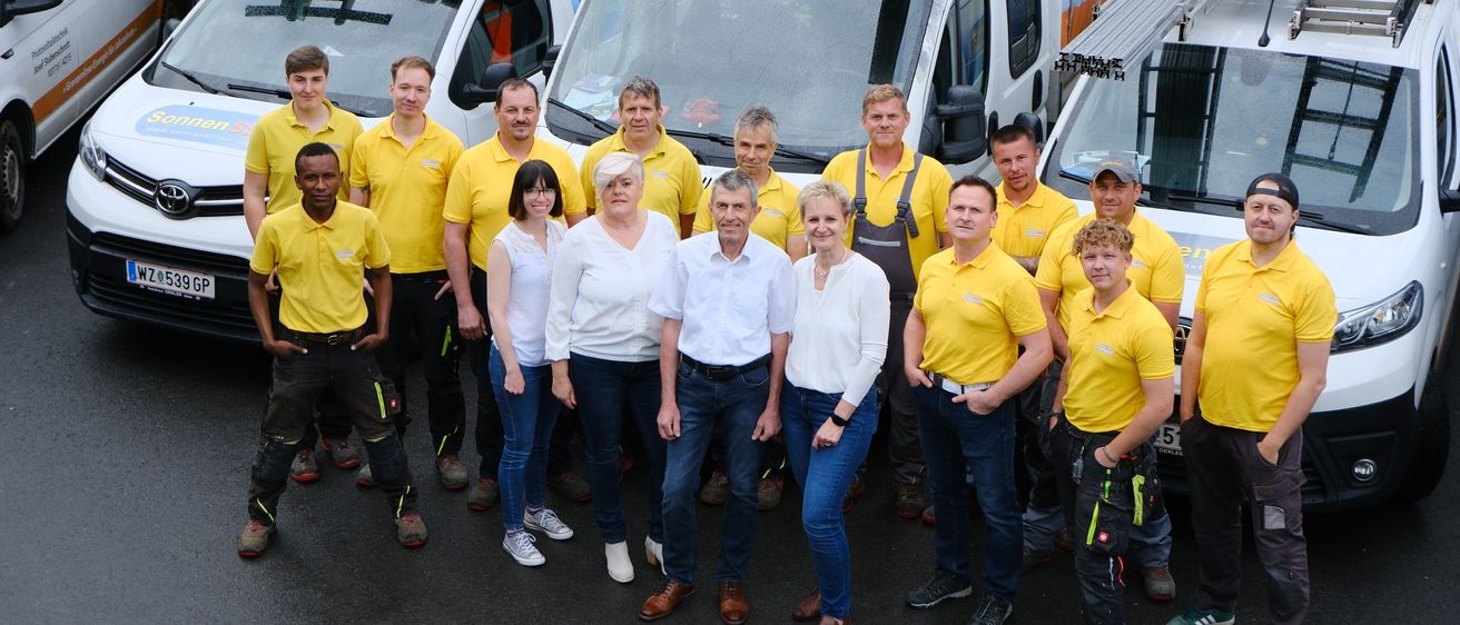 Eine Gruppe von Mitarbeitern in gelben Hemden steht vor drei weißen Lieferwagen, wobei zwei Männer in weißen Hemden und eine Frau in einem weißen Langarmshirt in der Mitte stehen.