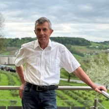 Ein älterer Mann steht auf einem Balkon mit Blick auf eine ländliche Landschaft. Der Hintergrund zeigt eine bewölkte Himmelslandschaft und grüne Hügel.