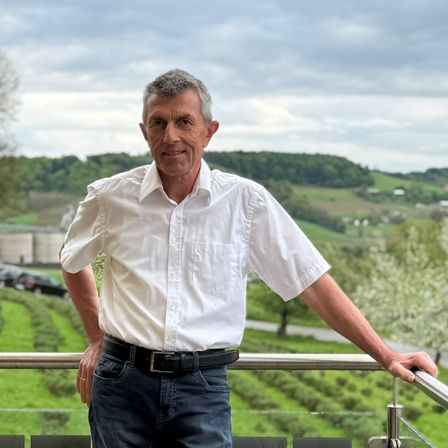 Ein älterer Mann steht auf einem Balkon mit Blick auf eine ländliche Landschaft. Der Hintergrund zeigt eine bewölkte Himmelslandschaft und grüne Hügel.