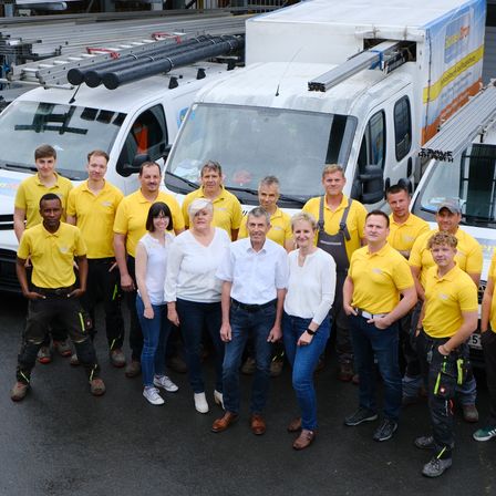 Eine Gruppe von Menschen in gelben Hemden posiert für ein Foto vor einer Reihe weißer Lieferwagen. Dahinter befinden sich Leitern und eine Metallkonstruktion.