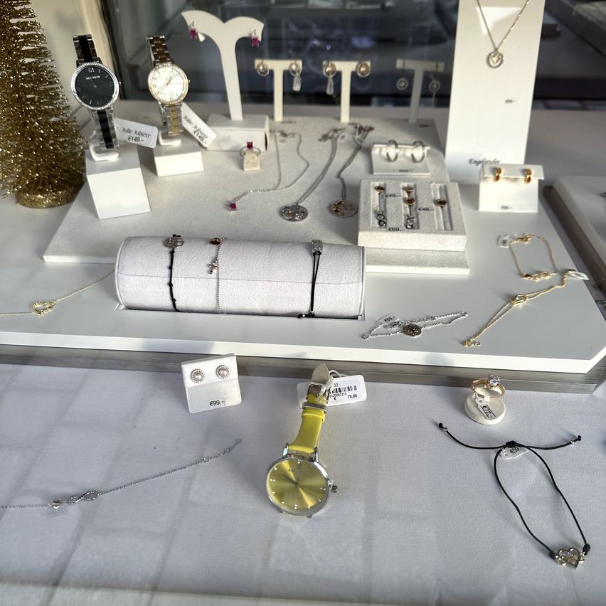 Ein weißer Tisch zeigt eine Schmuckkollektion. Es enthält Halsketten, Armbänder und Uhren. Einige Artikel haben Preisschilder. Ein Weihnachtsbaum steht auf der linken Seite.