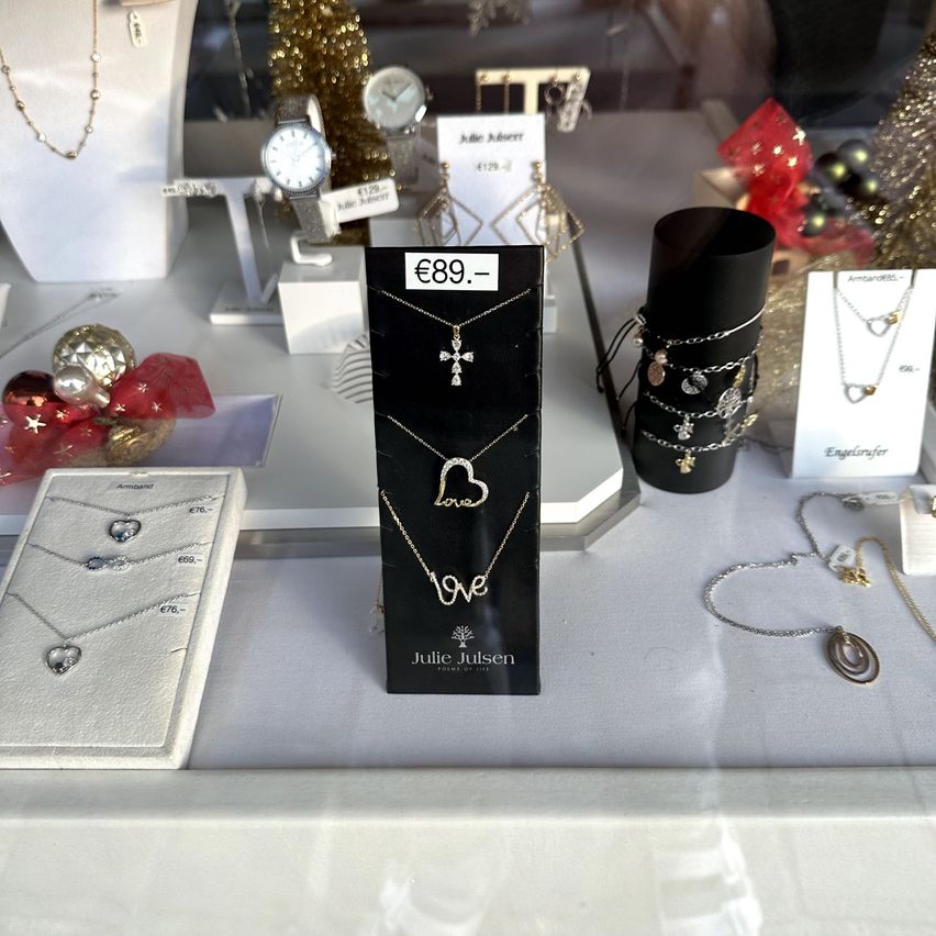 Eine Schmuckvitrine mit verschiedenen Halsketten, darunter herzförmige und Kreuzanhänger, auf einem weißen Tisch mit Preisschildern. Die Umgebung umfasst andere Schmuckstücke, Uhren und Weihnachtsdekorationen.