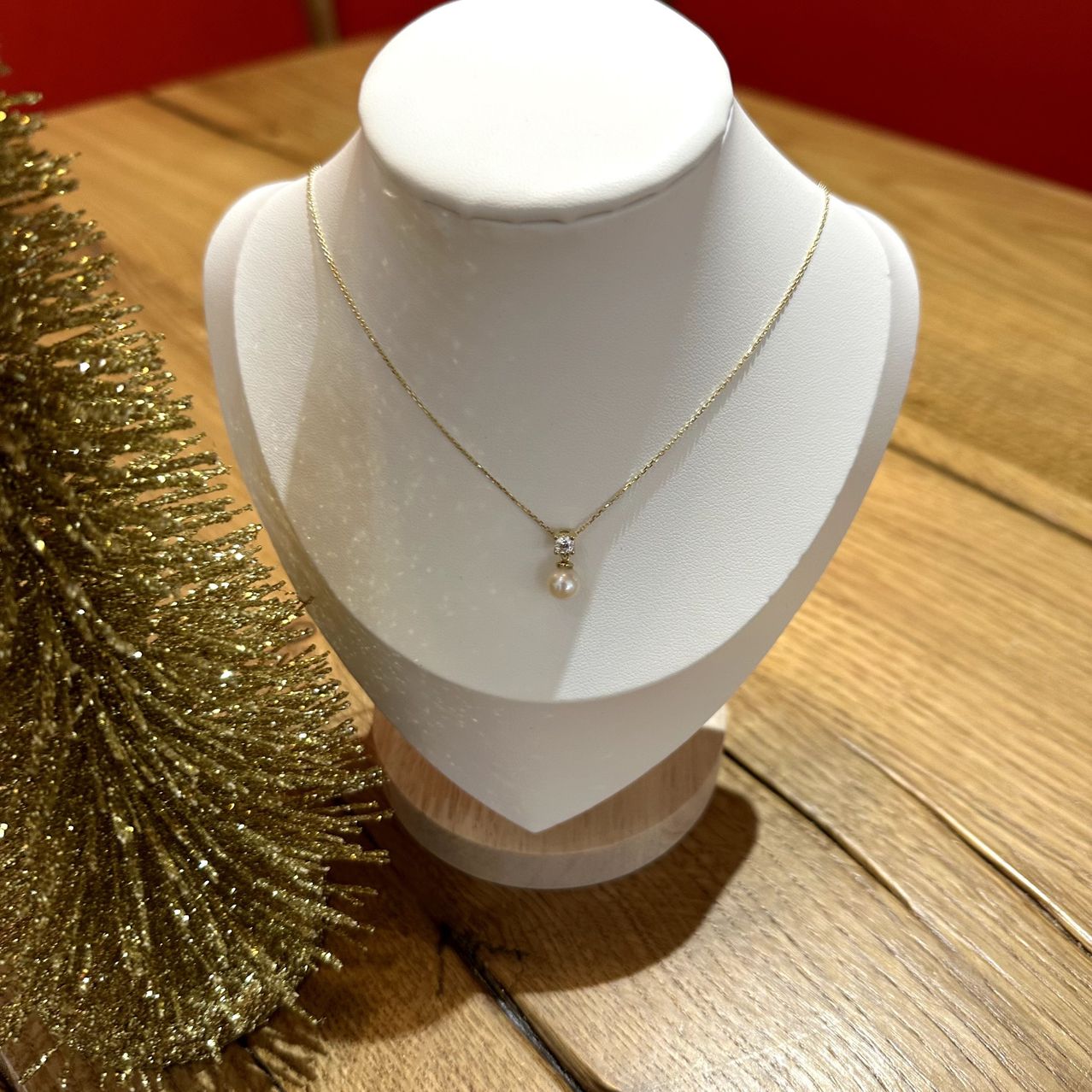 Eine goldene Halskette mit einem einzelnen Perlenanhänger wird auf einem weißen Ständer vor einem hölzernen Hintergrund neben einem goldenen Weihnachtsbaum ausgestellt.