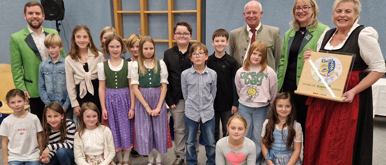 Eine Gruppe von Kindern und Erwachsenen posiert für ein Foto. Die Kinder tragen verschiedene Kleidungsstücke, und einige von ihnen halten ein Zertifikat. Die Erwachsenen stehen hinter ihnen, und einer von ihnen hält ein Zertifikat. Der Hintergrund ist eine blaue Wand mit einer Holzstruktur.