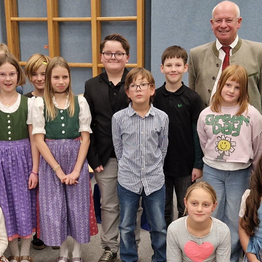Eine Gruppe von Kindern und Erwachsenen posiert für ein Foto. Die Kinder tragen verschiedene Kleidungsstücke, und einige von ihnen halten ein Zertifikat. Die Erwachsenen stehen hinter ihnen, und einer von ihnen hält ein Zertifikat. Der Hintergrund ist eine blaue Wand mit einer Holzstruktur.