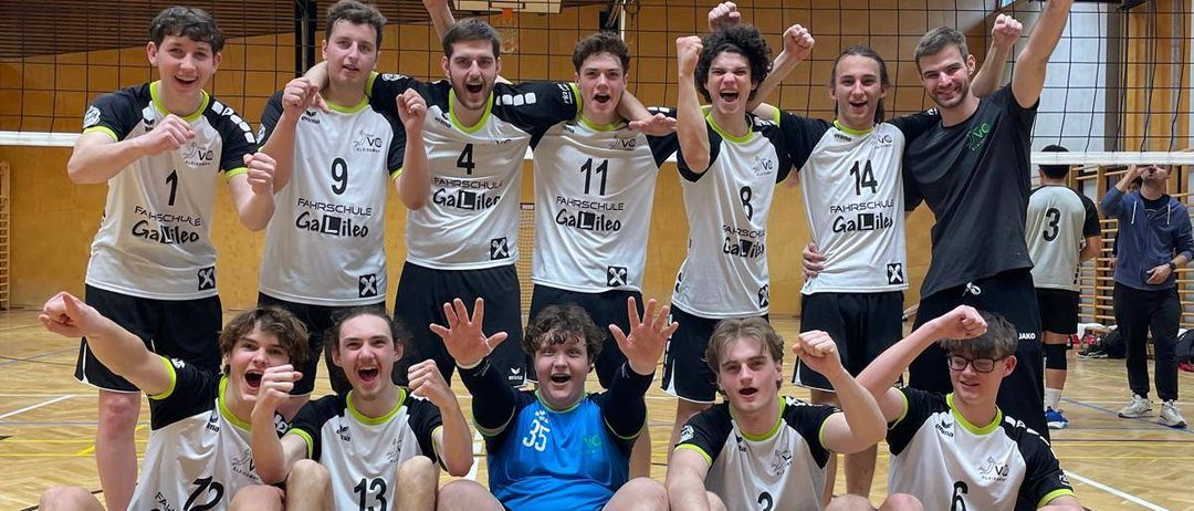 Eine Gruppe Volleyballspieler feiert gemeinsam auf einem Hallenplatz. Die Teammitglieder tragen weiße Trikots mit Nummern und einige tragen Brillen.