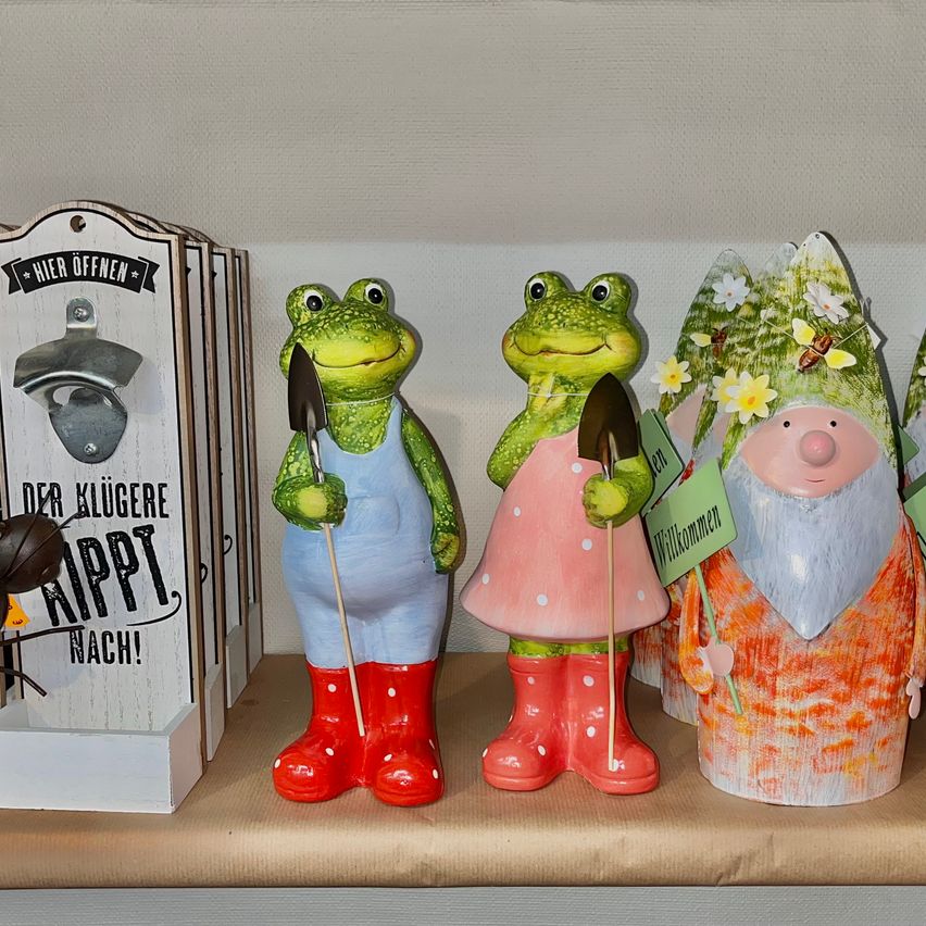 Vier Keramikfiguren sind auf einem Regal ausgestellt. Zwei Frösche halten Schaufeln, einer trägt Stiefel und der andere ein Kleid. Ein Kobold hält ein Schild. Dahinter befindet sich ein Flaschenöffner.