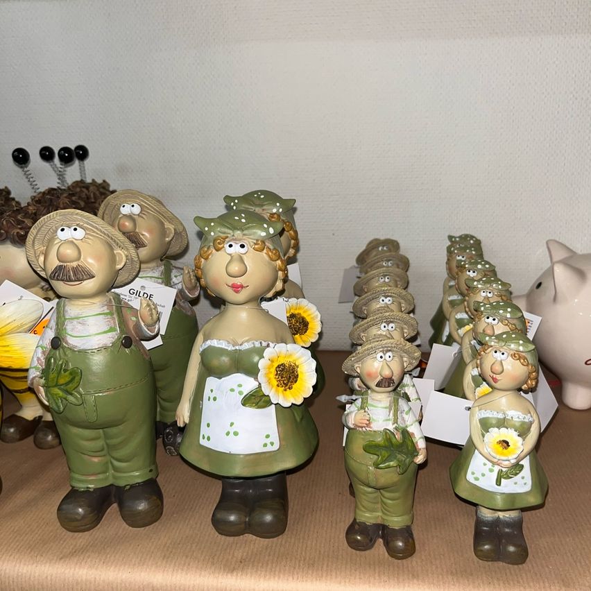 Eine Sammlung von Figuren, darunter Bauern mit Sonnenblumen, auf einer braunen Oberfläche. Einige Figuren haben Namensschilder, und im Hintergrund steht eine weiße Sparschweinbank.