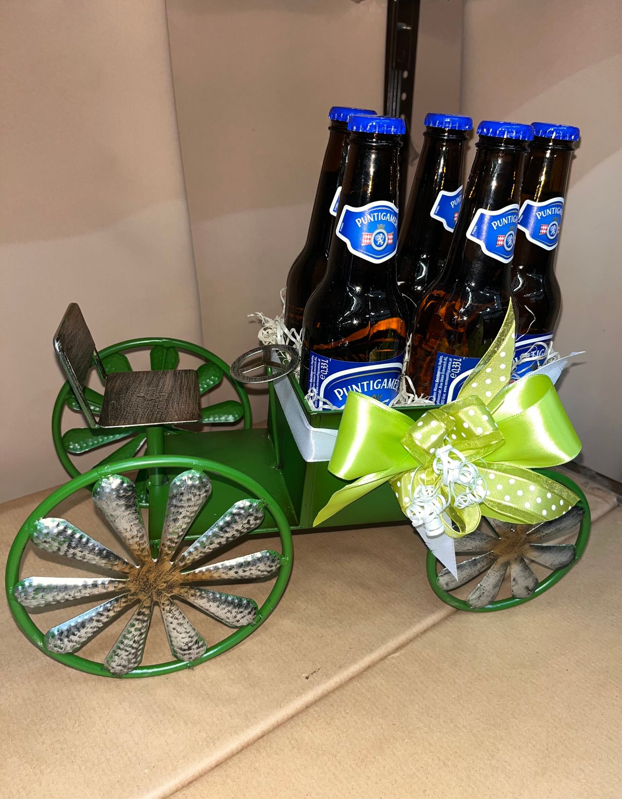 Ein grüner Wagen in Form eines Gespanns mit einem kleinen Sitz und Metallrädern, der sechs Puntigam-Bierflaschen enthält, mit einem grünen Band umwickelt.