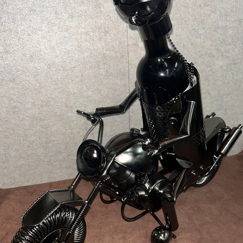 A black metallic sculpture of a motorcycle with a bottle as the rider is placed on a brown surface. The bottle is held by a chain and the sculpture has a shiny finish.