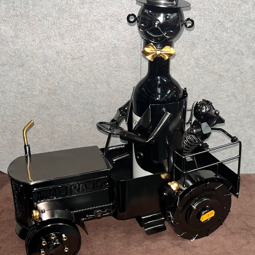 A black tractor-like object with a wine bottle head, gold bow tie, and dog figure in the back, on a brown surface.