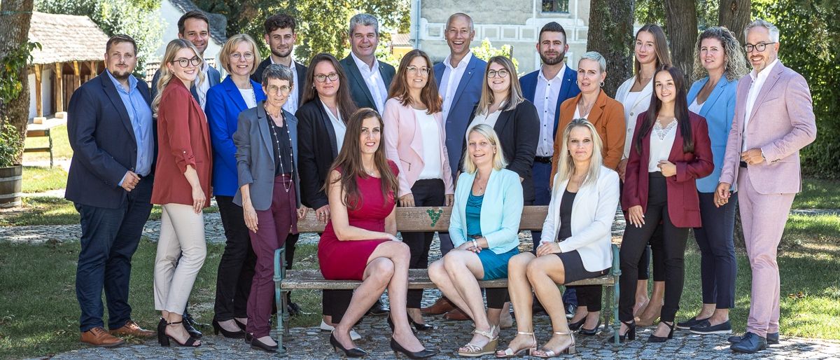 Eine Gruppe von Menschen in formeller Kleidung posiert für ein Foto im Freien. Sie sitzen auf einer Bank und stehen dahinter. Einige von ihnen tragen Brillen. Ein Baum steht hinter ihnen.