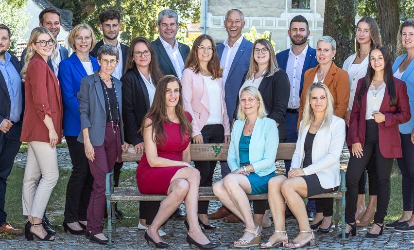 Eine Gruppe von Menschen in formeller Kleidung posiert für ein Foto im Freien. Sie sitzen auf einer Bank und stehen dahinter. Einige von ihnen tragen Brillen. Ein Baum steht hinter ihnen.