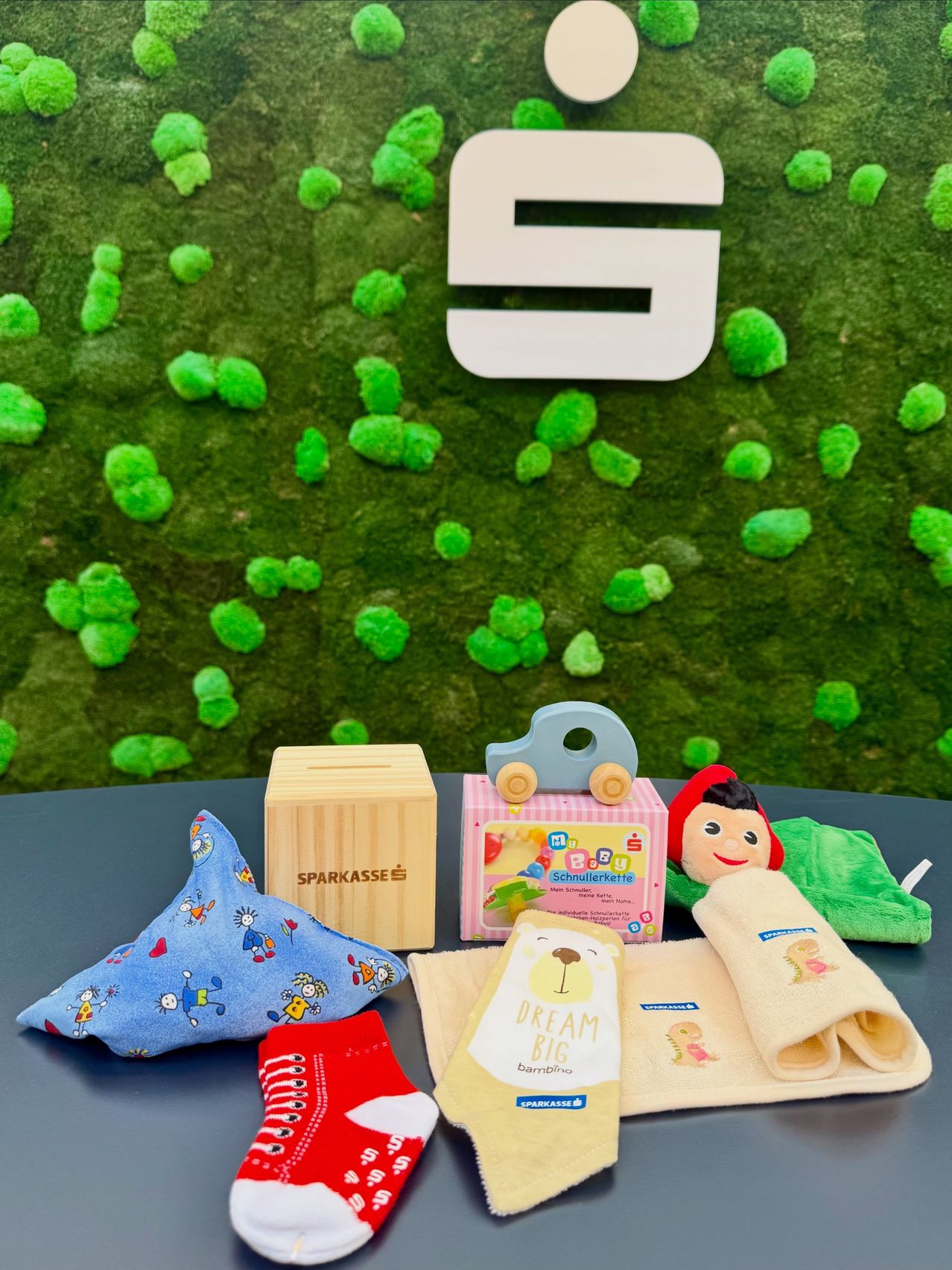 A collection of baby items on a table against a green moss wall with a large letter S. Items include a blue pillow, a wooden savings box, a pink box, a toy car, a doll, and a blanket.
