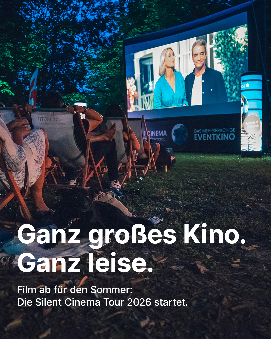 Menschen schauen draußen einen Film auf einer großen Leinwand. Der Film zeigt zwei Schauspieler vor einem Haus. Es ist eine Veranstaltung des ruhigen Kinos.