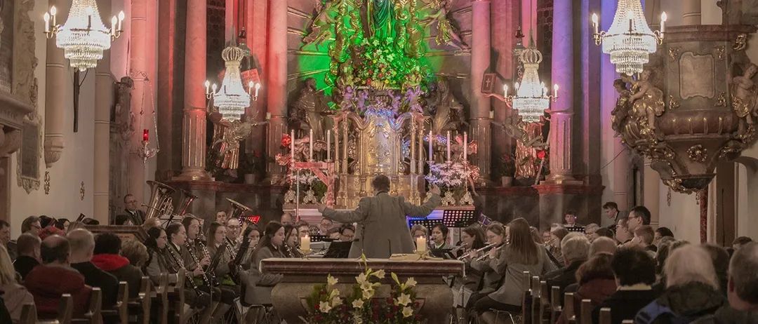 Ein Chor tritt in einer Kirche mit einem geschmückten Altar und beleuchtetem Hintergrund auf. Dirigent führt mit erhobenen Armen.