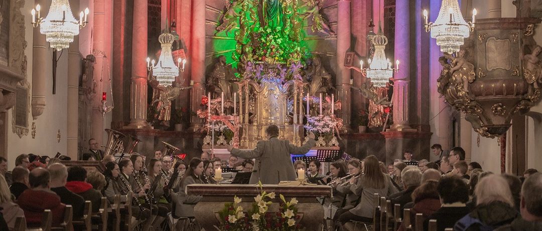 Ein Chor tritt in einer Kirche mit einem geschmückten Altar und beleuchtetem Hintergrund auf. Dirigent führt mit erhobenen Armen.
