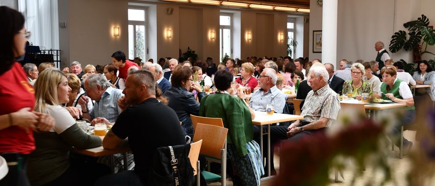 Viele Menschen sitzen um Tische in einem Speisesaal. Einige trinken aus Gläsern. Der Raum hat Fenster und Lampen.