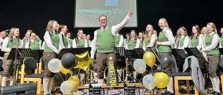 Eine Gruppe von Musikern und ein Dirigent stehen lächelnd auf der Bühne, mit Musikinstrumenten und Luftballons. Dahinter zeigt eine Projektionsleinwand eine Menschenmenge.