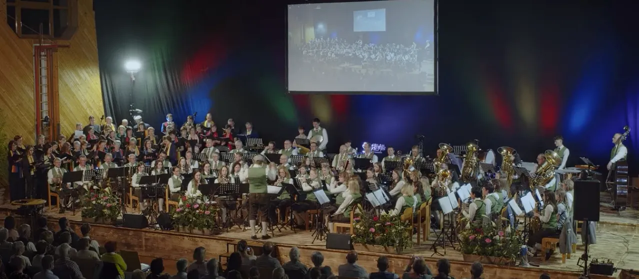 Ein Orchester auf der Bühne spielt Instrumente unter einer großen Leinwand. Die Musiker tragen grüne Westen und stehen zwischen Notenständern. Das Publikum sitzt unten.