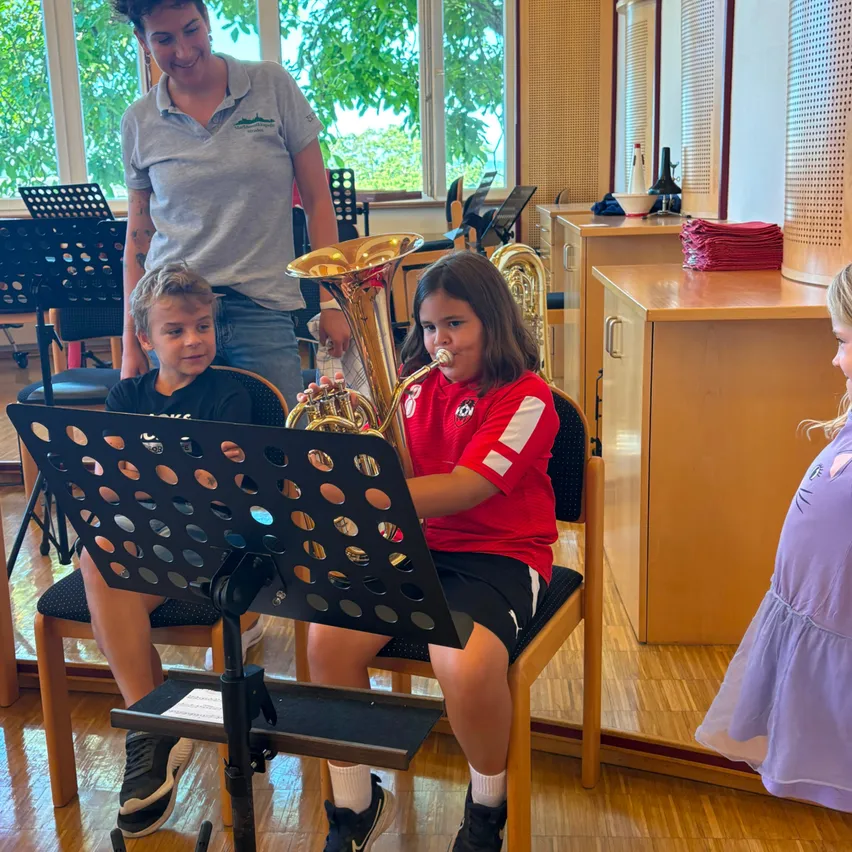 Zwei Kinder, eines spielt Tuba und das andere schaut zu, mit einer Frau, die hinter ihnen in einem Raum mit Musikinstrumenten und einem Fenster steht.