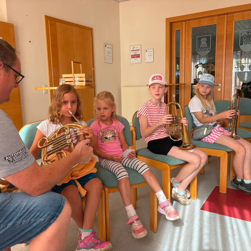 Ein Mann unterrichtet vier junge Mädchen, wie sie Blechblasinstrumente in einem Raum mit Holztüren und einer weißen Wand spielen.